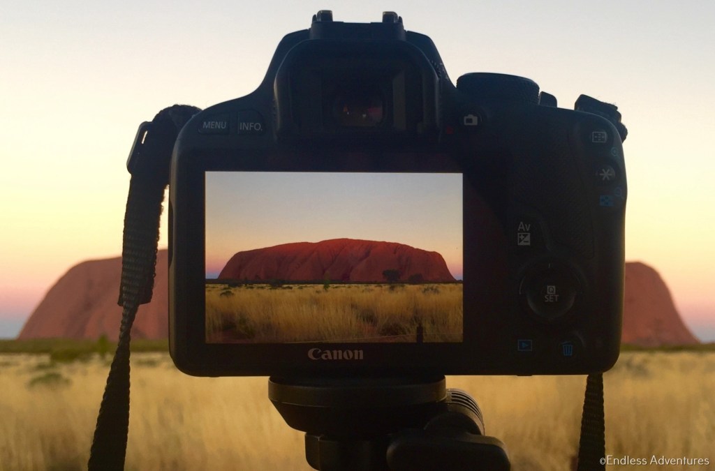 Camera on a tripod framing Uluru at sunset, with the rock visible on the camera screen