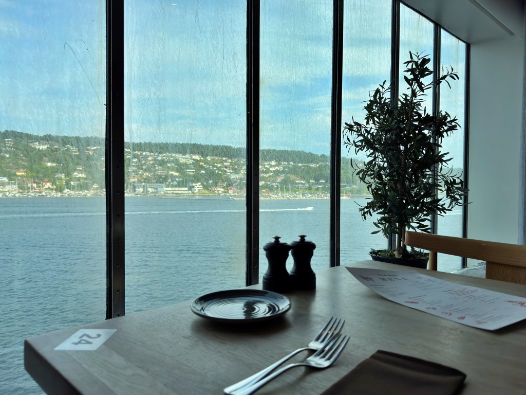 Table set for dining on a ferry, with cutlery, menu, and a view of the coastline through large windows