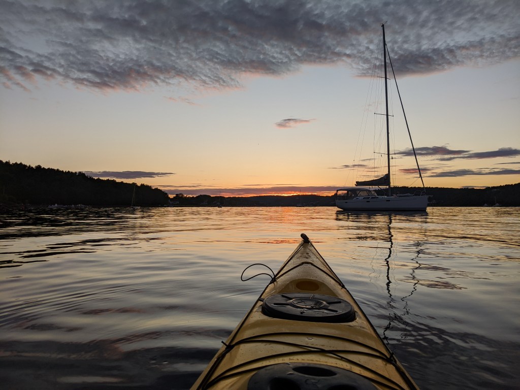 View from a kayak on calm water at sunset, with a sailboat nearby