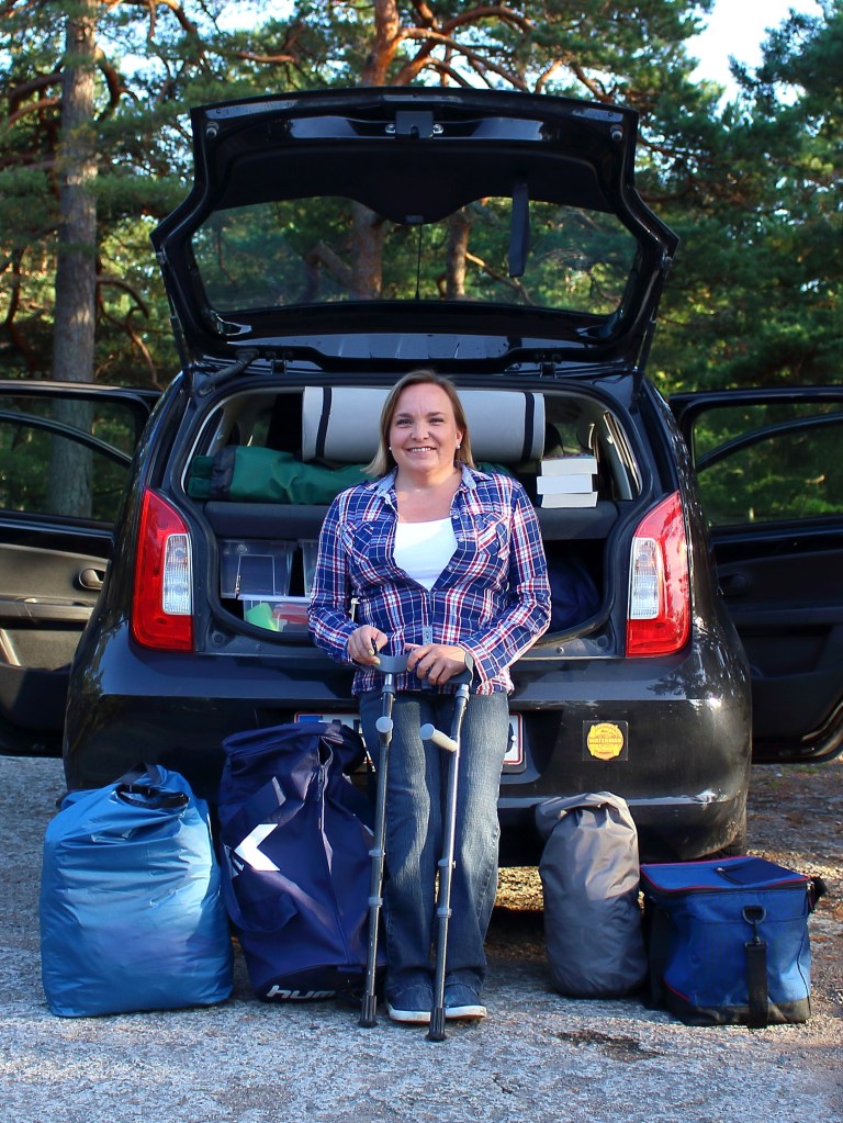 Adele Heidenreich standing on crutches beside an open car boot packed for a road trip